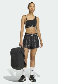 Reggiseno sportivo nero con una spallina, pantaloncini neri con dettagli lucidi, zaino nero, calzini bianchi con logo e scarpe da ginnastica nere.