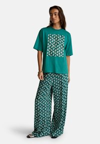 Tyrkysové bavlněné tričko s velkým vzorovaným čtvercem, spárované s kalhotami se širokými nohavicemi s opakujícím se vzorem listů v krémové barvě na tyrkysovém pozadí.