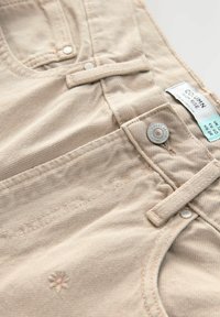 Pantalons beiges taille haute en tissu texturé, dotés d'une fermeture par bouton visible, de passants de ceinture et de détails de broderie florale subtile.