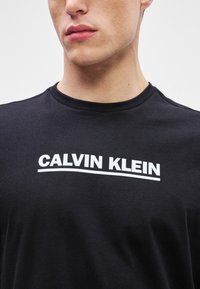 T-shirt de algodão preto com decote redondo, apresentando o logo em branco "CALVIN KLEIN" em destaque no peito. Textura suave, corte casual.
