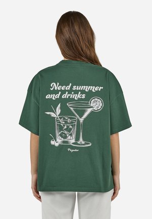Person mit langen braunen Haaren, der ein grünes T-Shirt mit weißen Aufdrucken von Cocktailgläsern und dem Text trägt: "Brauche Sommer und Drinks."