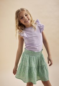 Lila t-shirt med spetsiga volanger ärmar, i kombination med en grön blommig lagerkjol. Kjolen har ett smockat midjeband och en utfläkt form.