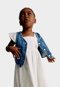 Džínová vesta s špičatým límcem a knoflíky, nošená přes bílé šaty s krátkými rukávy. Dítě stojí sebevědomě s rukama v bok.