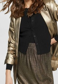 Metalen gouden blazer, zwart geribd vest met knopen en geplooide bronzen rok met glanzende textuur. Close-up van de kledingdetails.