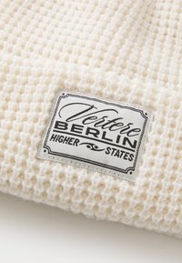 Beanie lavorato a maglia color crema con un motivo testurizzato e un'etichetta cucita che reca la scritta "Vertere Berlin Higher States" in nero e grigio.
