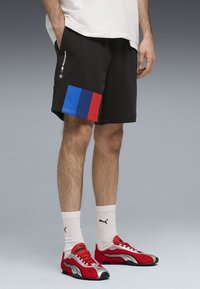 Shorts de survêtement noirs avec des accents de bandes rouges et bleues à l'ourlet, dotés de poches latérales et d'un logo sur le devant. Portés avec des chaussures de sport rouges.