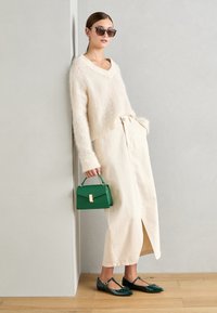 Light beige oversized sweater with a v-neck, cream pleated midi skirt, green leather handbag, and glossy green flats with bows.