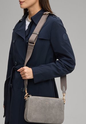 Vrouw in een marineblauwe trenchcoat met een grijze suède crossbodytas met brede verstelbare band en goudkleurige details.
