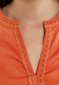 Blusa arancione con scollatura a V presenta cuciture decorative a scalloped lungo i bordi e una texture di tessuto liscia.