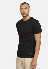 T-shirt noir à manches courtes, col rond, texture côtelée, coupe ajustée, avec un petit logo près de l'ourlet. Stylisé avec un pantalon de couleur claire.