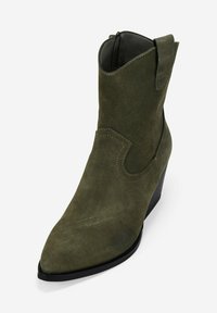 Botín de ante verde oliva hasta el tobillo con punta afilada, tirador, cremallera lateral, costuras decorativas y tacón en cuña apilada.