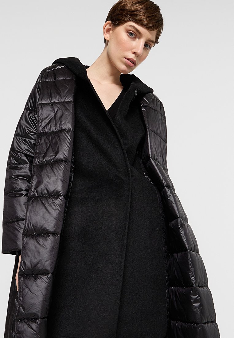 Czarna kurtka puchowa o pikowanej fakturze i kapturze, noszona na gładkiej czarnej sukience z wełny. Materiał wydaje się gładki i miękki.