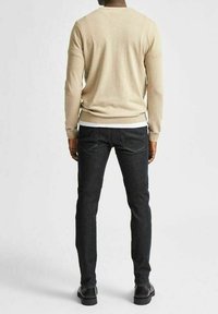 Pull beige en maille douce, avec col rond et poignets côtelés, associé à un jean foncé ajusté avec coutures jaunes. Chaussures noires.