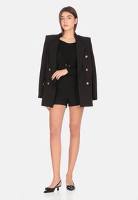 Blazer nero con bottoni dorati, indossato sopra un top e pantaloni corti neri. Tacchi a punta neri con dettagli floreali. Tessuto liscio, vestibilità su misura.
