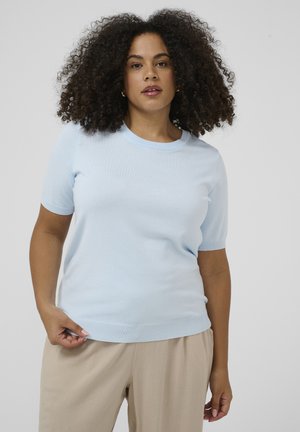 Frau mit lockigem Haar, die einen hellblauen Kurzarmpullover und beige Hose trägt, steht vor einem schlichten hellgrauen Hintergrund.