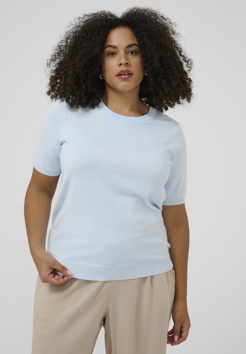 Femme aux cheveux bouclés, portant un pull léger à manches courtes bleu clair et un pantalon beige, debout devant un fond gris clair uni.