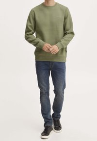 Felpa verde oliva con scollatura rotonda e polsini, abbinata a jeans blu e sneakers nere, su uno sfondo neutro.