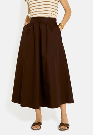 Persona con un top de rayas beige y marrón, falda midi marrón de talle alto, sandalias marrones y una pulsera dorada, con las manos en los bolsillos de la falda.