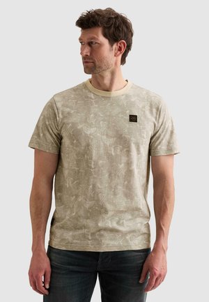 Homme aux cheveux courts et bouclés, portant un t-shirt beige à manches courtes avec un motif et un jean foncé, regardant sur sa gauche sur un fond uni.