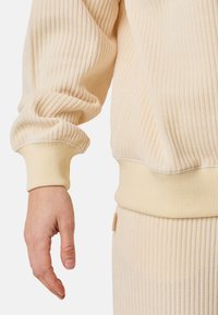 Beige geribde trui met een ontspannen pasvorm, met een omgeslagen zoom en mouwen, gestructureerde stof en een subtiele glans. Hand ligt ernaast.
