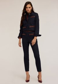 Costume en denim bleu marine avec une veste courte mettant en valeur des accents et des boutons en cuir marron, associé à un pantalon slim et des escarpins bordeaux.