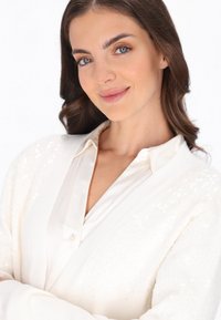 Weißer Pailletten-Cardigan über einer cremefarbenen Satinbluse mit Kragen und Knopfdetails. Weiche Textur, lässige Passform und sanfter Glanz.