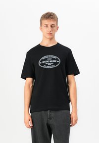 Jaunas vyras stovi tiesiai, vilkėdamas juodą marškinėlį su baltu apvaliu tekstu „Jack & Jones Worldwide“ ir tamsiai pilkomis džinsinėmis kelnėmis.