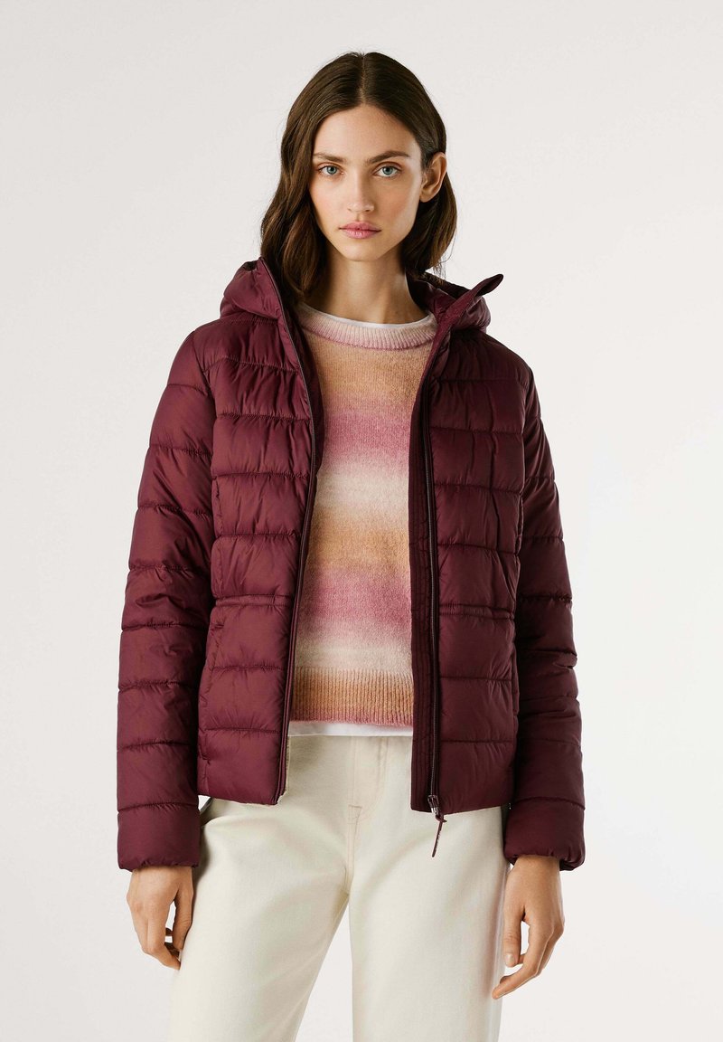 Vrouw in een donkerrode gewatteerde jas over een veelkleurige roze en beige trui met crèmekleurige broek, staand tegen een effen achtergrond.