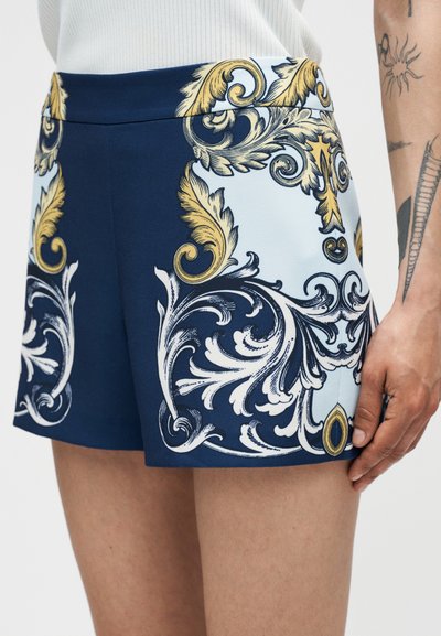 Μπλε και ανοιχτό μπλε σορτς με χρυσά και λευκά μπαρόκ σχέδια που φορά ένα άτομο με τατουάζ στο εμφανές χέρι και μπράτσο.