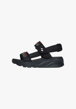 Sandalias negras con correas de tela que presentan acentos verticales coloridos. Suela gruesa y texturizada con un diseño curvado y correa ajustable en el talón.