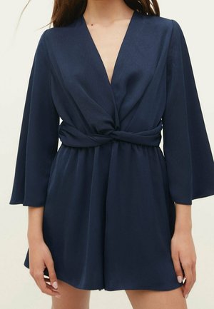Vrouw die een marineblauw playsuit met lange mouwen draagt, met een diepe V-hals en een gedraaide knoop in de taille.