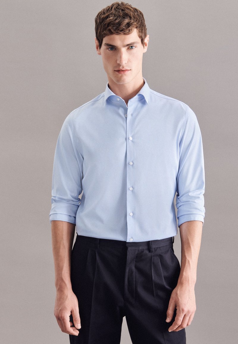 Camicia button-up azzurro chiaro con maniche arrotolate, caratterizzata da un colletto classico e una texture morbida, abbinata a pantaloni scuri plissettati.