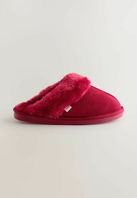 Piros papucs puha szövetből, bolyhos, puha mandzsettával. Ívelt formájú, lapos és masszív talppal rendelkezik. Látható márkajelzés.