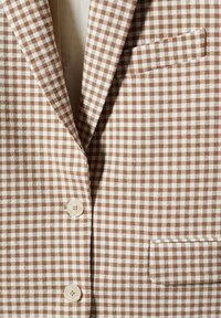 Blazer à motif vichy marron et blanc, avec un revers cranté, deux boutons et un détail de poche poitrine. La texture du tissu semble tissée.