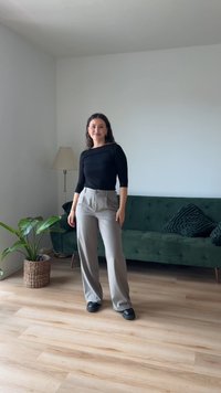 Camisa preta de mangas longas e calças largas cinza claro. O conjunto é complementado com sapatos pretos. Sofá verde e planta ao fundo.