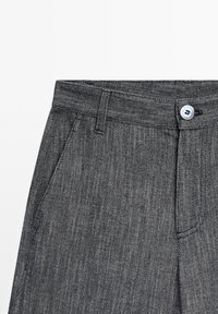 Grossissement d’un pantalon gris texturé avec poche avant, passant de ceinture et bouton blanc à la taille, sur fond blanc.