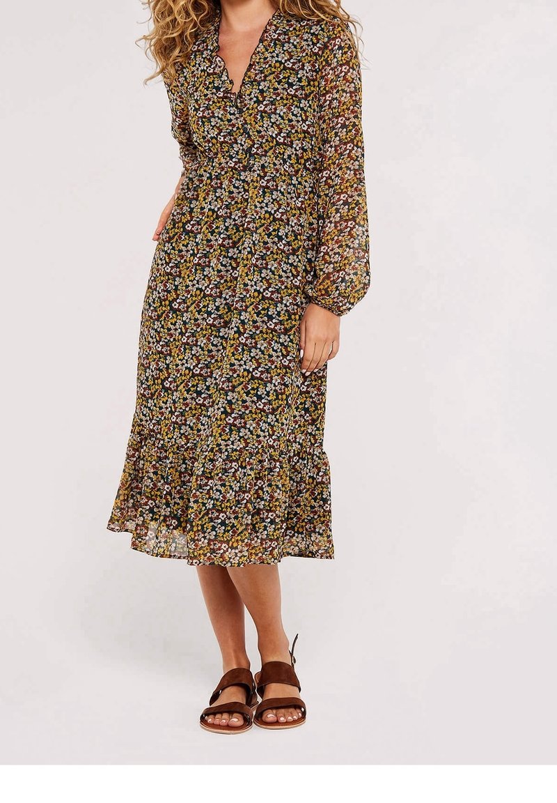 Kobieta w długiej sukience midi w kwiatowe wzory z falbaną na dole, nosząca brązowe, otwarte sandały na płaskim obcasie.