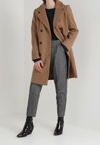 Kamelfärgad dubbelknäppt kappa över en mörkgrön ribbad tröja, i kombination med grå mönstrade byxor och blanka svarta ankelstövlar.