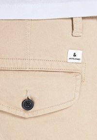 Poche de pantalon en tissu beige avec un bouton noir et une petite étiquette de marque blanche cousue près de la passants de ceinture.