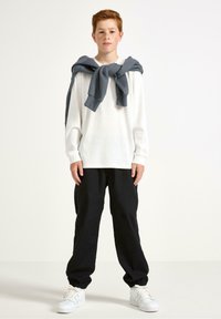 Ragazzo dai capelli rossi che indossa una camicia bianca a maniche lunghe con un maglione grigio legato sulle spalle, pantaloni neri e sneakers bianche, in piedi contro uno sfondo bianco.
