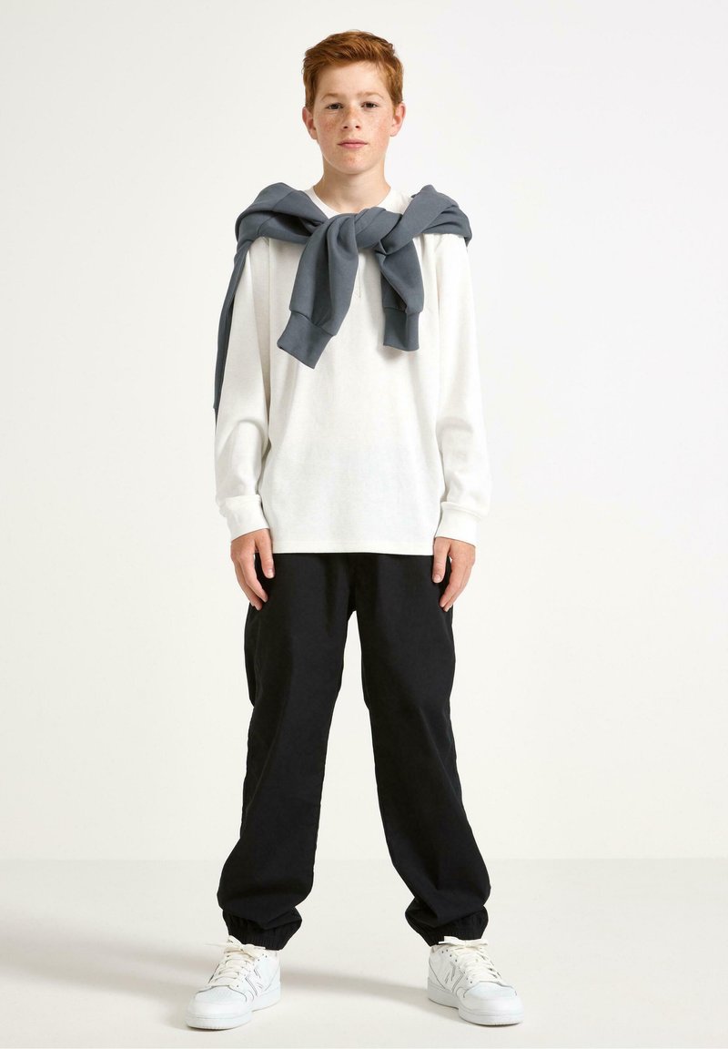 Ragazzo dai capelli rossi che indossa una camicia bianca a maniche lunghe con un maglione grigio legato sulle spalle, pantaloni neri e sneakers bianche, in piedi contro uno sfondo bianco.