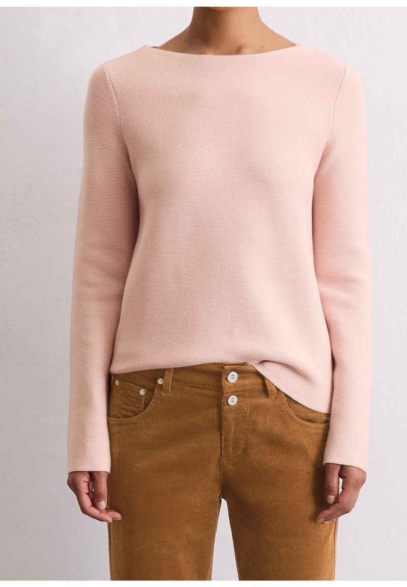 Personne portant un haut en maille à manches longues rose clair rentré dans un pantalon en velours côtelé marron taille haute avec double boutons.