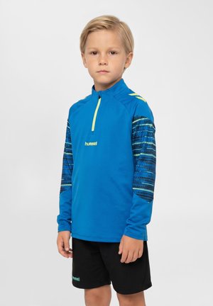 Jeune garçon debout avec une expression neutre, portant un pull de sport bleu avec des manches à motifs et un short noir.