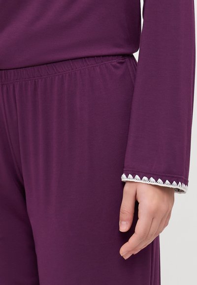 Top lung cu mâneci lungi de culoare mov, cu o margine texturată albă la mansete, asortat cu pantaloni cu talie elastică. Material moale, ușor.