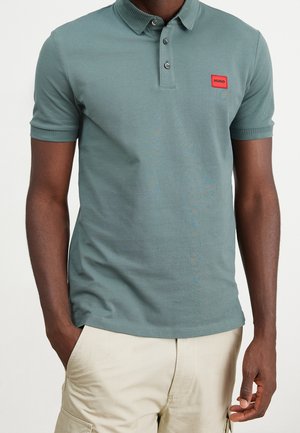 Mann trägt ein grünes Polohemd mit kurzen Ärmeln mit rotem Hugo-Logo-Aufnäher und hellbeige Cargo-Shorts, Hände in den Taschen.