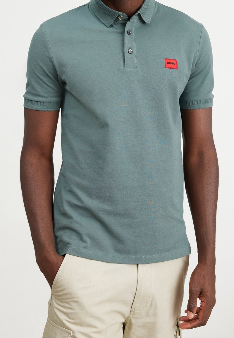 Homme portant un polo vert à manches courtes avec un patch logo Hugo rouge et un short cargo beige clair, mains dans les poches.