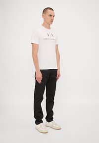 Hvit T-skjorte med "A|X ARMANI EXCHANGE" logo, svarte bukser og beige sneakers med minimalistisk design og teksturert stoff. Stående mannlig figur.