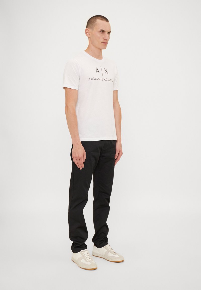 Hvit T-skjorte med "A|X ARMANI EXCHANGE" logo, svarte bukser og beige sneakers med minimalistisk design og teksturert stoff. Stående mannlig figur.