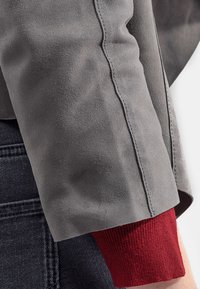 Grijze suede jacksleeve met een zichtbare naad, voorzien van een rode geribbelde manchette. De achtergrond toont een donkere spijkerstoftextuur.