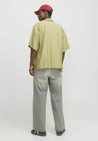 Kurzärmliges, gelbes Musterhemd kombiniert mit hellgrauen, weiten Jeans und einer roten Baseballmütze, von hinten betrachtet. Einfache Designdetails sichtbar.